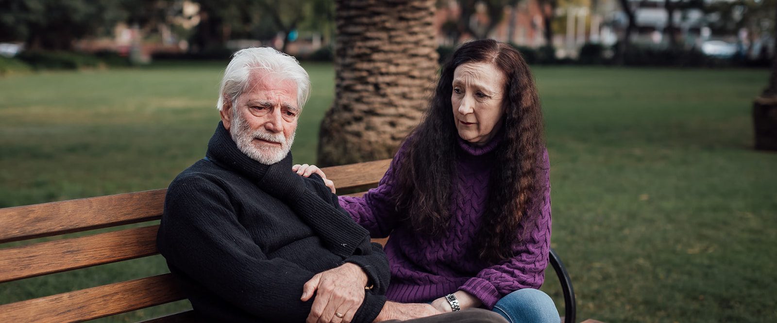 An upset older man sitting on a park bench being comforted by a woman with long dark hair.