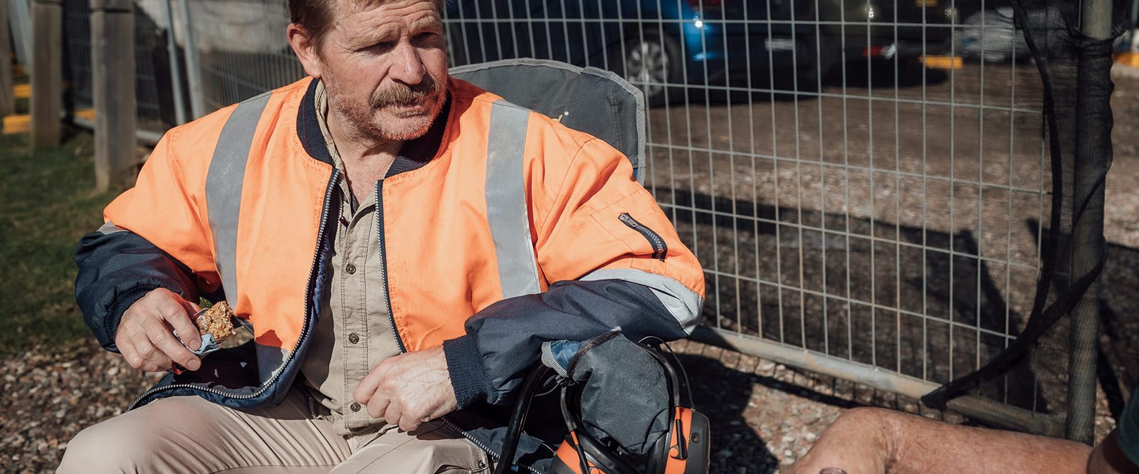 An unshaven man in a hi vis jacket and brown pants, sitting talking to another man.