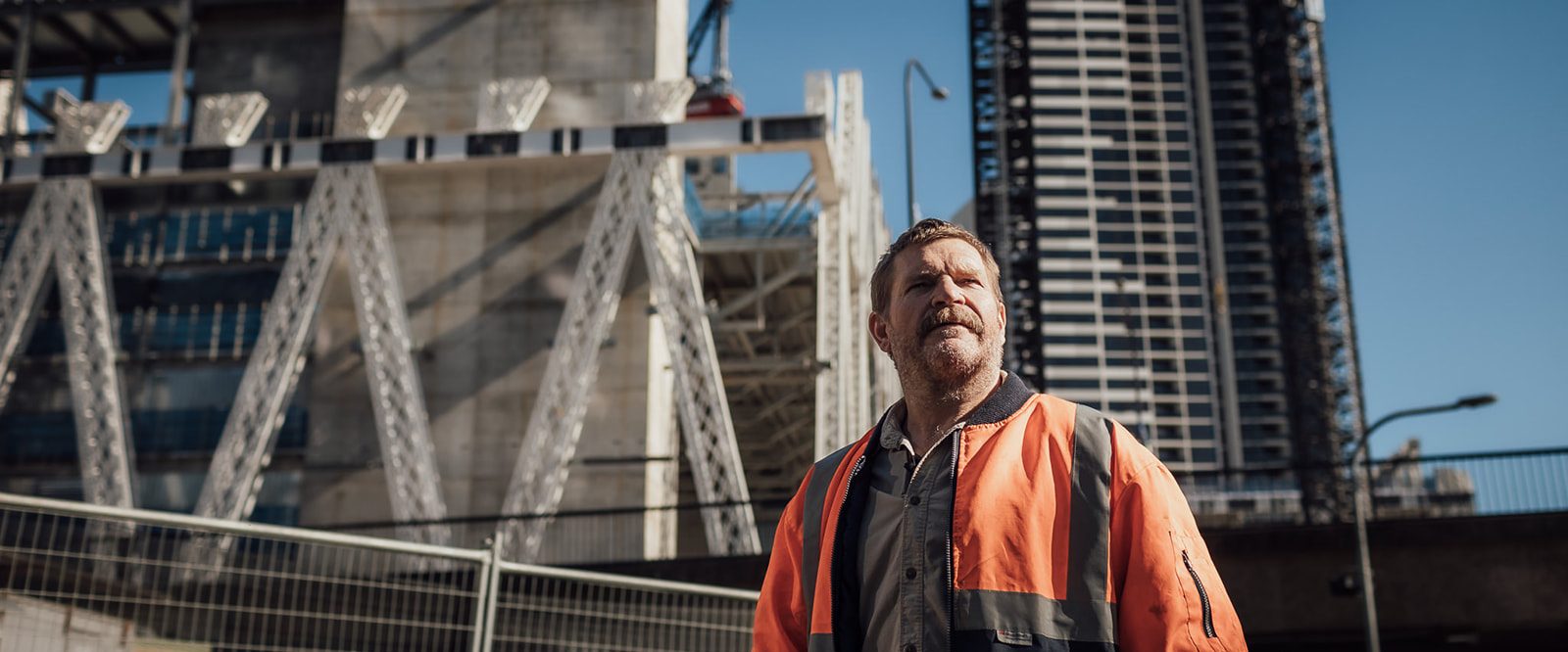 A man with a moustache and unshaven face wearing a high vis jacket, looking thoughtfully standing in front of a building under construction.
