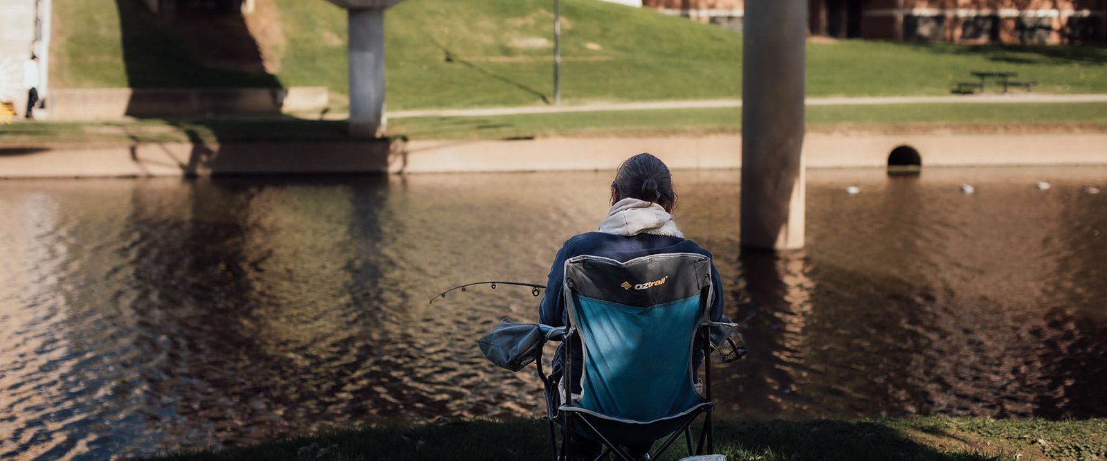The back of a man seated, fishing by the river.
