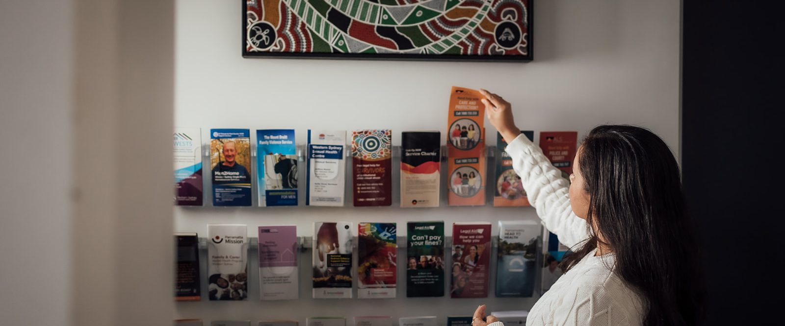 A female selecting Aboriginal brochures from a brochure wall underneath an Aboriginal painting.