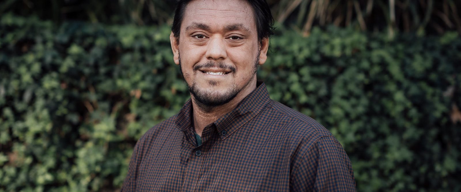 A smiling man wearing a brown business shirt, standing in front of a leafy hedge.
