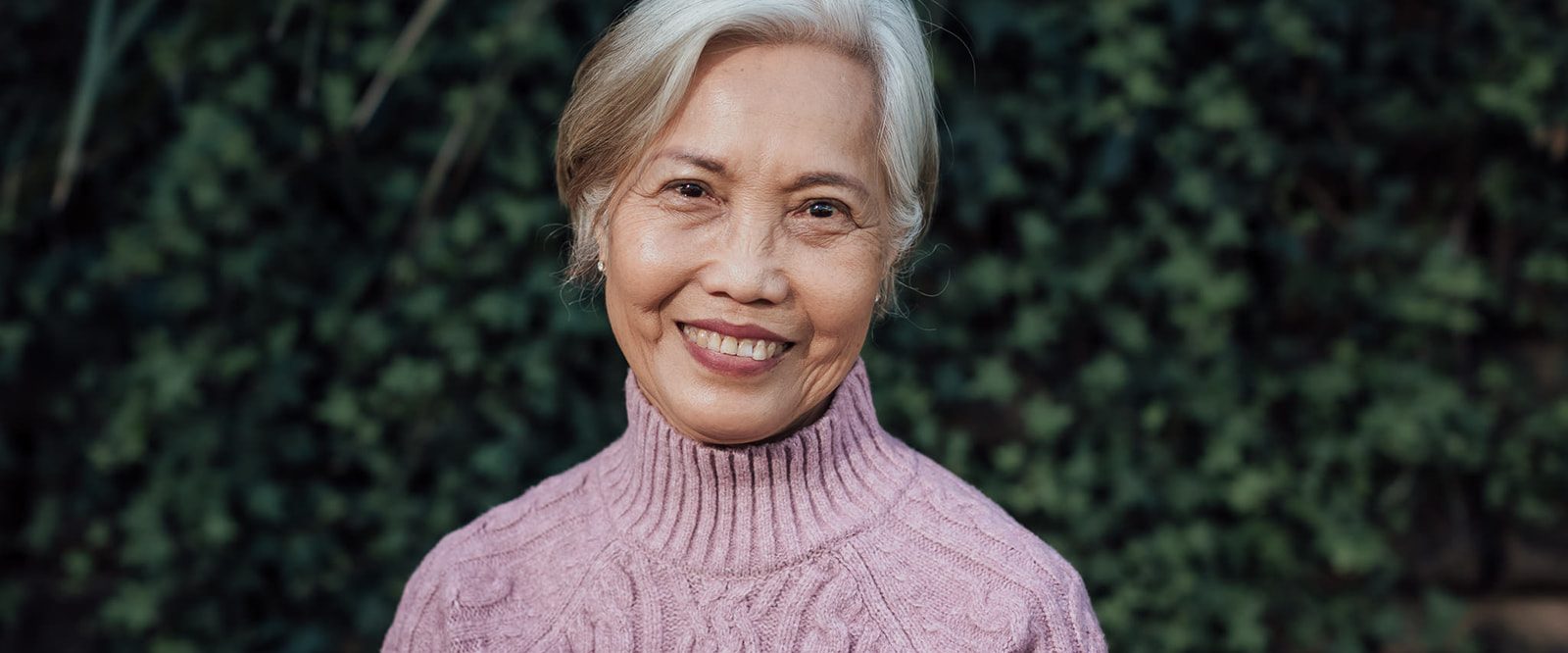 An older lady smiling, wearing a pink jumper, standing in front of a leafy wall.