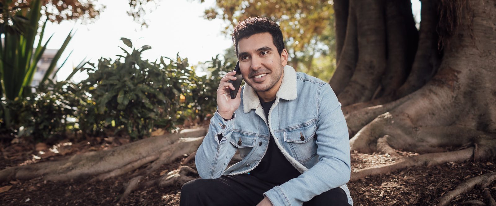 A smiling man sitting underneath a tree talking on the phone.
